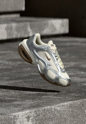 Baskets de sport crème et argent métallique flottant au-dessus de marches en béton, avec semelles en caoutchouc marron et logo Nike.