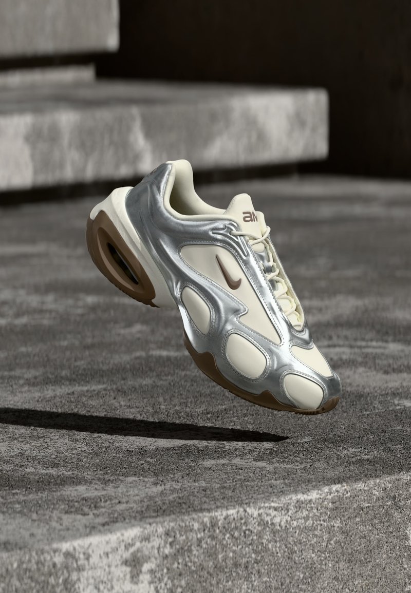 Αθλητικό παπούτσι σε κρεμ και μεταλλικό ασημί χρώμα, που αιωρείται πάνω από σκάλες από μπετόν, με καφέ λαστιχένιες σόλες και το λογότυπο της Nike.