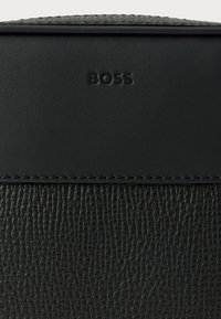 Μαύρη δερμάτινη τσάντα με υφή στο κάτω μέρος και λείο πάνω μέρος, με ανάγλυφο λογότυπο "BOSS" και αντίθετη ραφή.