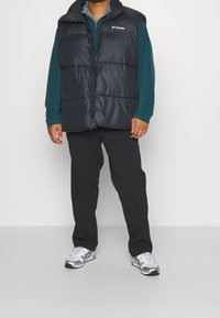 Chaleco acolchado azul marino con cuello alto, cierre frontal y logotipo bordado. Llevado sobre una camiseta de manga larga color aqua y combinado con pantalones negros y zapatillas.