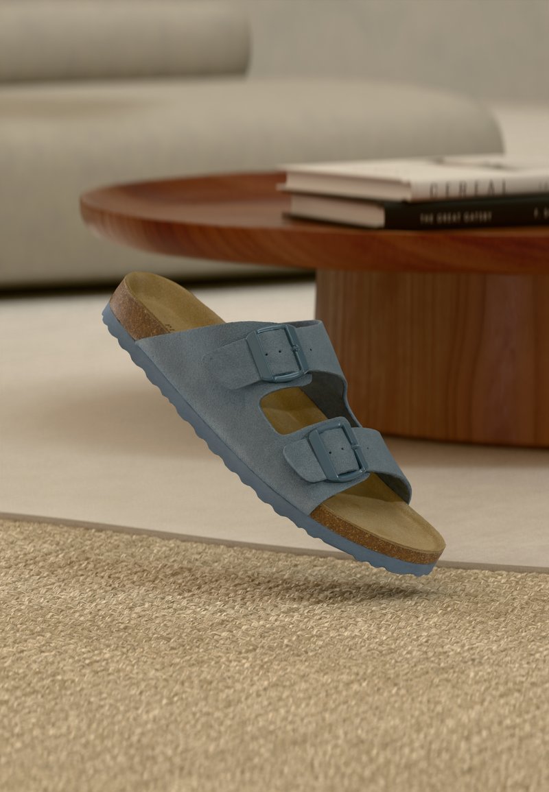 Sandale en daim bleu avec deux boucles réglables, semelle intérieure en liège et semelle texturée, flottant au-dessus d'un tapis beige tissé dans un salon.