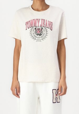 Persoon die een lichtbeige Tommy Jeans T-shirt draagt met een roze tijgerlogo en witte broek met een grote "H" patch op het dijbeen.
