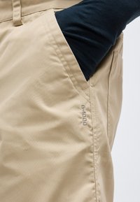 Μπεζ παντελόνι από ύφασμα G-1000 με πλαϊνή τσέπη, μαύρο μανίκι που φαίνεται μερικώς μέσα στην τσέπη, κοντινή λήψη στη ραφή και την υφή του υφάσματος.