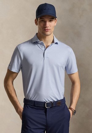 Férfi világoskék mintás galléros pólót, sötétkék nadrágot övvel, sötétkék sapkát és karórát visel, kezét zsebében tartva áll.