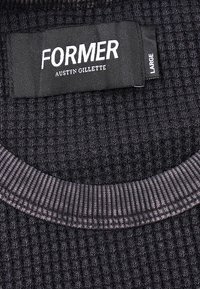 Μαύρο υφαντό ύφασμα με ανάγλυφη υφή, με ριμπ λαιμόκοψη και μαύρη ετικέτα που γράφει "FORMER Austyn Gillette" και ετικέτα μεγέθους "Large" σε μικρότερη πλαϊνή ετικέτα.