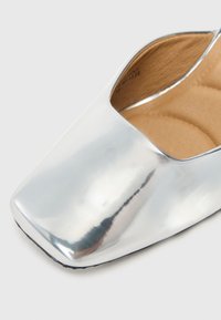 Μοναδικό ασημί μεταλλικό φλατ παπούτσι με τετράγωνη μύτη και καφέ μαλακή εσωτερική επένδυση σε ουδέτερο φόντο.