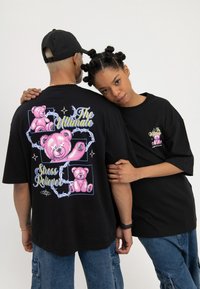 Două persoane purtând tricouri negre supradimensionate cu desene de ursuleți de pluș roz și textul „The Ultimate Stress Reliever”, una cu fața spre înainte, cealaltă cu spatele.