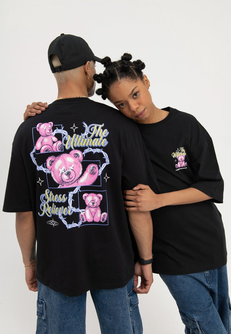 Două persoane purtând tricouri negre supradimensionate cu desene de ursuleți de pluș roz și textul „The Ultimate Stress Reliever”, una cu fața spre înainte, cealaltă cu spatele.
