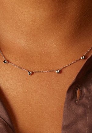 Zilveren hartvormige hangersketting met vijf kleine hartaccenten aan een delicate ketting, rustend op gebruinde huid, minimalistisch ontwerp.