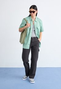 Camisa de botão de manga curta verde claro, t-shirt cinza, calças escuras riscas de giz, sapatilhas cinzas, a carregar uma mala tote verde, a usar óculos de sol.