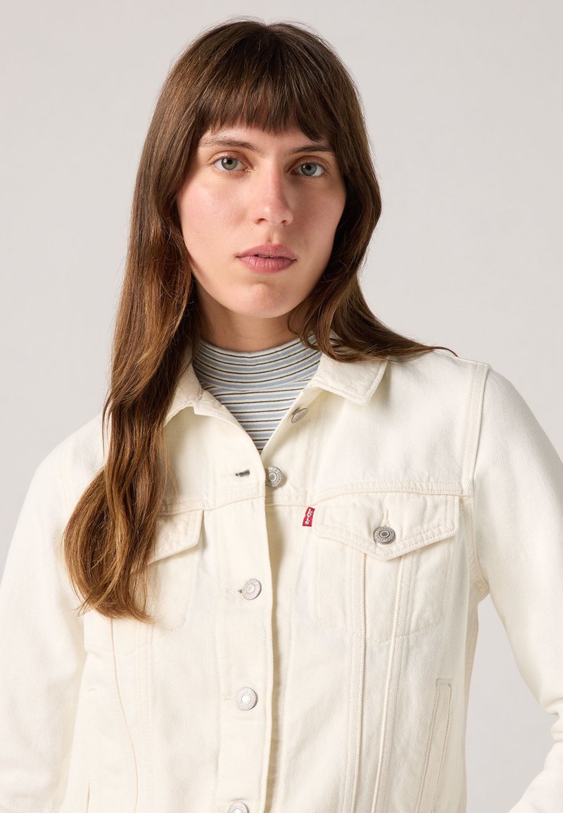 Chaqueta de mezclilla blanca con dos bolsillos en el pecho, cierre de botones y etiqueta roja de la marca. Llevada sobre un suéter de cuello alto a rayas gris y blanco.