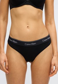 Mustad puuvillased püksikud, millel on vormiv disain ja pehme vöökoht, kus on valge kirjaga "Calvin Klein" logo. Sile tekstuur, minimaalne detail.