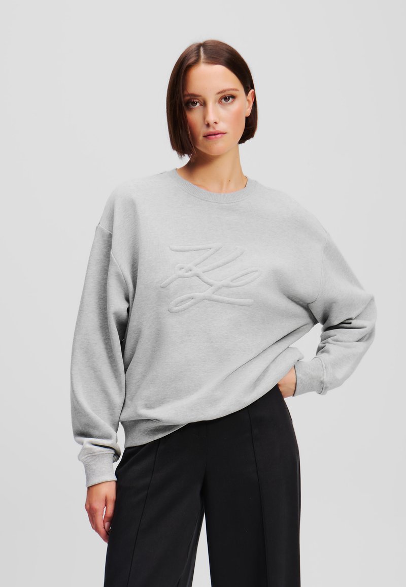 Felpa grigia chiara con un logo ricamato testurizzato sul davanti. Presenta un collo a giro e una vestibilità oversize, realizzata in un tessuto morbido.