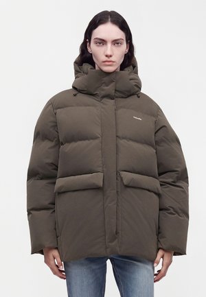 Personne aux cheveux raides et foncés portant une doudoune oversize vert olive foncé et un jean bleu, debout devant un fond uni.