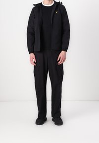 Zwarte capuchonjack met een ritssluiting aan de voorkant, gecombineerd met een zwart T-shirt met ronde hals en zwarte cargobroek met meerdere zakken, en zwarte schoenen.