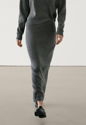 Person iført en tætsiddende grå strikket sweater og matchende lang nederdel, går indendørs på glat gulv med sorte lædersko.