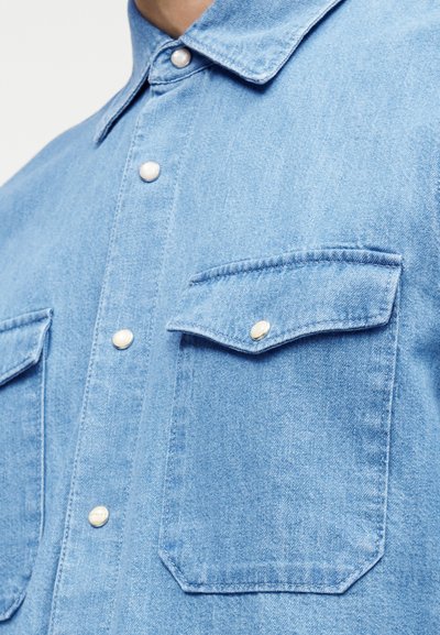 Light blue denim shirt with pearl snap buttons and two chest pockets, one with a flap and button closure.