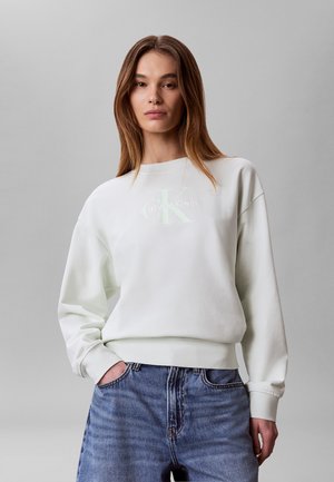 Kobieta o prostych brązowych włosach, ubrana w biały sweter Calvin Klein oraz niebieskie dżinsy, stojąca na tle jednolitego szarego tła.