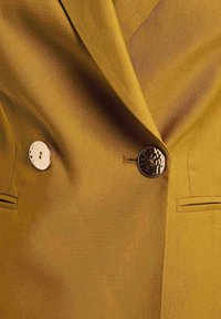 Blazer amarillo con una tela texturada, dos botones distintivos—uno metálico y otro con un patrón—junto a un bolsillo en el lado izquierdo.