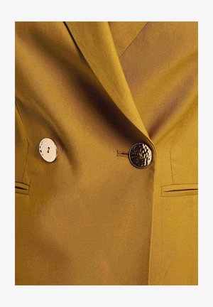 Geel blazer van een geaccentueerde stof, met twee onderscheidende knopen - een metalen en een met een patroon - naast een zak aan de linkerkant.