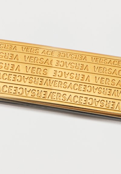 Cinci bare de aur paralele, cu cuvântul "VERSACE" înscris în relief de mai multe ori pe toată lungimea lor pe un fundal alb.