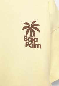 Keltainen puuvillapaita, jossa on ruskea palmupuu-grafiikka ja "Baja Palm" -teksti alla. Lyhyet hihat, klassinen malli, pehmeä materiaali.