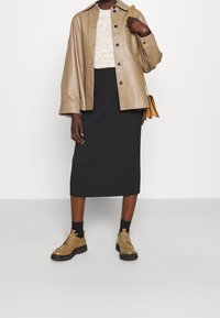 Veste en cuir tan avec des boutons, pull texturé clair, jupe noire côtelée mi-longue, chaussures tan avec semelles noires, et un petit sac jaune.