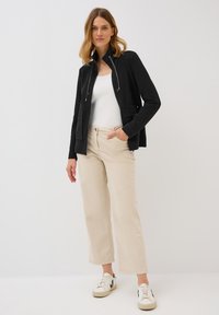 Eine Frau steht selbstbewusst in einer schwarzen Reißverschlussjacke, einem weißen Oberteil, beigen Hosen und Turnschuhen vor einem neutralen Hintergrund.