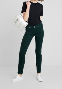 Gröna kordjeans i skinny-modell med mjuk textur och fem fickor, ihop med en svart t-shirt och vita sneakers. Inkluderar en vitväska.