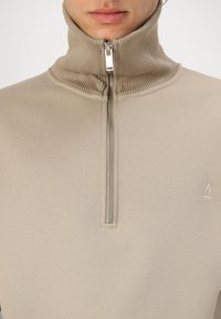 Suéter beige con cremallera y un alto cuello acanalado, cremallera plateada y un sutil detalle de logotipo. Tejido suave con un diseño ajustado.