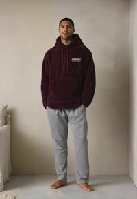 Vīnsarkanā fleeces džemperī ar priekšējā kabatu un baltā krāsā uzdrukātu uzrakstu "SNOOPY", kopā ar pelēkām sporta biksēm. Modelis stāv basām kājām pret vienkāršu sienu.