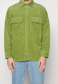 Camicia di velluto a coste verde con colletto, due tasche sul petto, chiusura frontale con bottoni e maniche lunghe, caratterizzata da una texture a coste verticali.