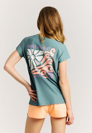 Vrouw met lang haar die een turquoise T-shirt draagt met de tekst "Comes in waves" en een golfontwerp, gecombineerd met perzikkleurige shorts, staand tegen een effen achtergrond.