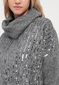 Grå strikkegenser med skulderhals, prydet med sølvfargede paljetter ordnet i vertikale striper, som tilfører tekstur og detaljer.