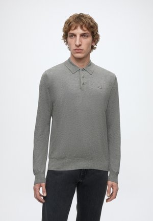 Junger Mann mit welligem Haar, der ein graues langärmeliges Poloshirt und schwarze Jeans vor einem schlichten weißen Hintergrund trägt.