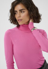 Pull ajusté à col roulé rose avec encolure côtelée, manches longues et texture en maille fine. Porté avec une ceinture noire et un bracelet métallique.