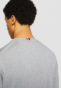 Uomo con capelli corti e ricci volto di spalle, che indossa un maglione grigio chiaro con dettagli a righe rosse, bianche e nere sul collo sul retro.