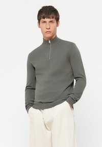 Grå genser med glidelås i en tettsittende design, laget av lett strikket stoff. Har høy krage og ribbestrikkede mansjetter. Brukt sammen med beige bukser.