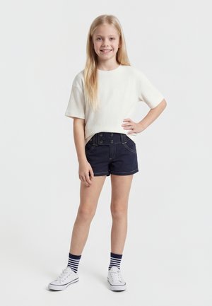 Mladé dievča sa usmieva, stojí s jednou rukou na páse, oblečená v bielom tričku s krátkym rukávom, námorníckych kraťasoch, pruhovaných ponožkách a bielych teniskách.