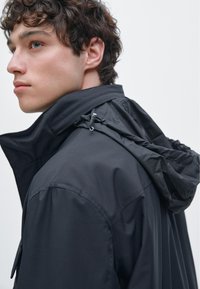 Heren zwarte waterdichte jas met een hoge kraag, verstelbare capuchon en ritssluitingen. Soepele textuur, minimalistisch ontwerp en praktische kenmerken.