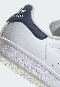 White sneaker featuring a textured leather upper, navy blue heel tab, and rubber sole with patterned traction for grip.