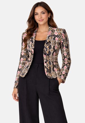 Mujer con un blazer estampado con diseños en rosa, beige y negro, sobre un top negro y pantalones anchos negros, con una mano en el bolsillo.