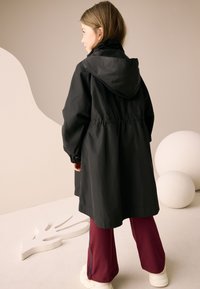 Veste noire imperméable avec capuche, taille cintrée et poignets élastiques, portée avec un pantalon bordeaux à bandes latérales bleues et des baskets blanches.