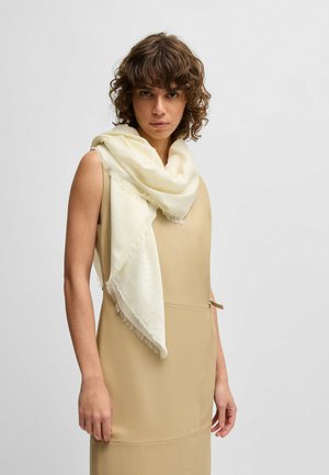 Bufanda blanca con bordes flecados colocada sobre un vestido beige. La bufanda tiene una textura suave y una apariencia ligeramente transparente.