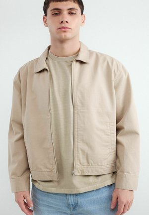 Młody mężczyzna w beżowej kurtce na zamek błyskawiczny, beżowej koszulce i jasnoniebieskich dżinsach, stojący na jednolitym białym tle.