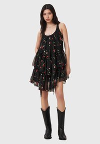 Schwarzes ärmelloses Kleid mit Schichten aus rüschigem, transparenter Stoff, verziert mit einem floralem Muster aus roten und weißen Blumen. Schwarze Cowboy-Stiefel.