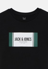 Melnā kokvilnas T-krekls ar taisnstūra logotipu, kurā ir teksts "JACK & JONES" baltā un zaļā krāsā, apaļš kakls, gluda tekstūra.