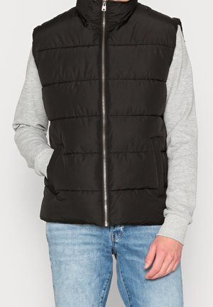 Muž nosící černou prošívanou vesta bez rukávů přes světle šedou mikinu a vybledlé modré džíny, s jednou rukou v kapse vesty.