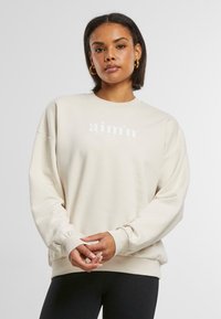 Cremefarbener Sweatshirt mit langen Ärmeln, gerippten Bündchen und rundem Halsausschnitt. Besitzt weißen Markentext auf der Vorderseite. Weicher Stoffeindruck.