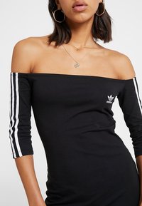Svart off-shoulder klänning med trekvartsärmar, med vita ränder längs ärmarna och en liten vit Adidas-logga.